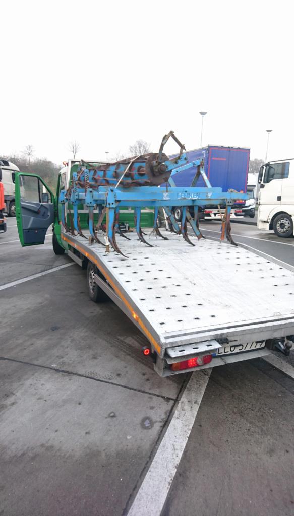 Zielona laweta z otwartymi drzwiami, przewożąca niebieski, metalowy kultywator rolniczy na perforowanej platformie, na parkingu z innymi pojazdami w tle.
