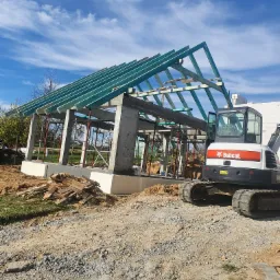 Wznoszenie szkieletu budynku z betonu i drewna z użyciem podpór, na placu budowy stoi minikoparka Bobcat, w tle błękitne niebo z chmurami.