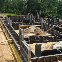 Szare metalowe szalunki fundamentowe z żółtymi elementami wzmacniającymi, częściowo wypełnione ziemią, na tle zieleni drzew i błękitnego nieba z chmurami.