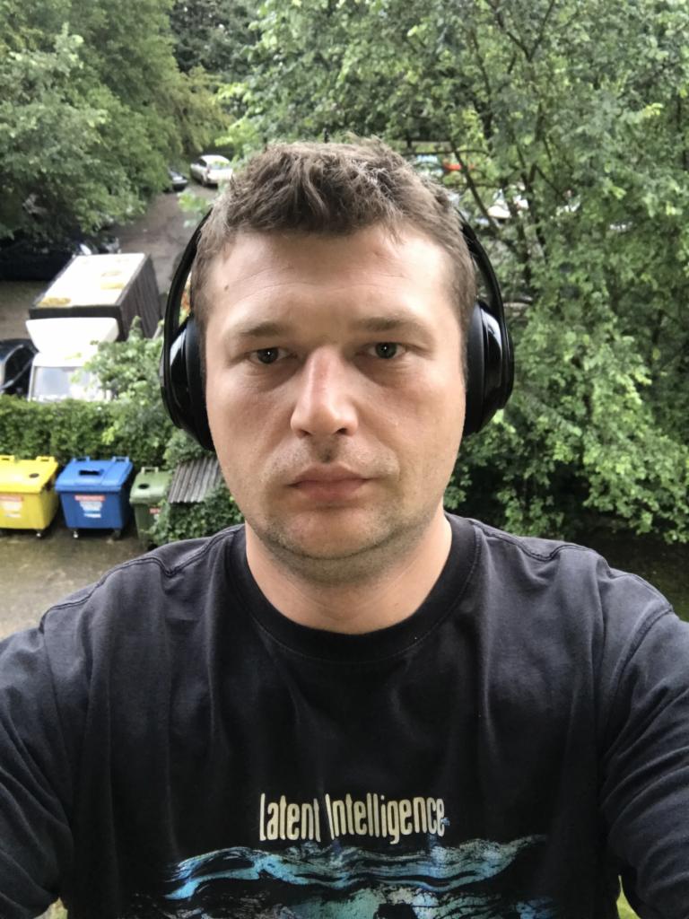 Mężczyzna w czarnej koszulce z napisem 'Latent Intelligence' i słuchawkach na uszach, z widokiem na drzewa i podwórze z kontenerami na śmieci w tle.