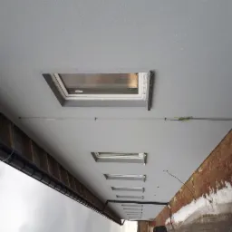 Szara elewacja budynku z oknami dachowymi i widoczną instalacją elektryczną na zewnątrz, z fragmentem ceglanej ściany po prawej stronie.