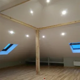 Poddasze z drewnianą konstrukcją szkieletową, oknami dachowymi i wykończoną podłogą imitującą drewno, przygotowane do dalszych prac wykończeniowych.