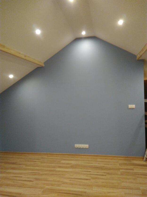 Poddasze po remoncie: ściana pomalowana na szaro-niebieski kolor, drewniana podłoga, widoczne punkty świetlne w suficie i fragment konstrukcji drewnianej.