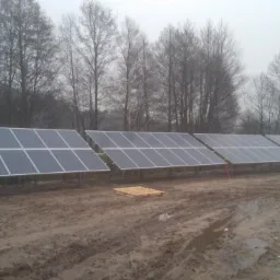 Rząd paneli słonecznych na metalowych konstrukcjach ustawiony na błotnistym terenie, w tle zalesiony obszar w mglisty dzień.