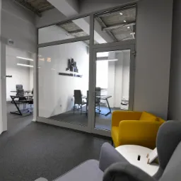 Nowoczesne wnętrze biurowe z przeszkloną ścianą działową, widoczne biurka i krzesła, żółty fotel w strefie relaksu, szara wykładzina dywanowa.
