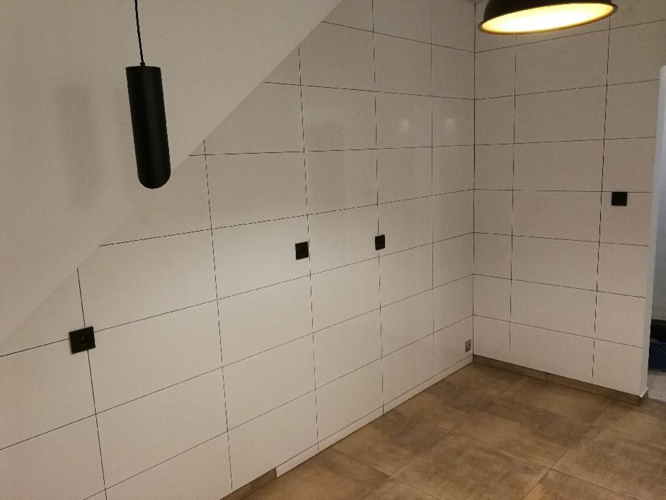 Ściana i podłoga wyłożone białymi płytkami z ciemnymi fugami, czarne punkty elektryczne, wisząca czarna lampa, brązowa podłoga, skos na ścianie.