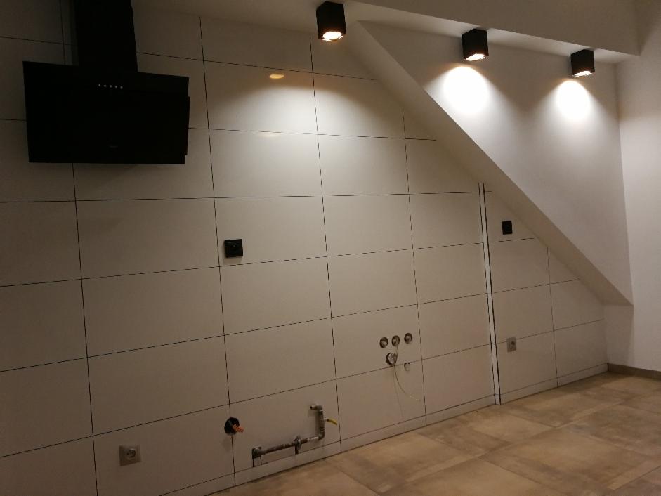 Ściana w kuchni wyłożona białymi płytami wielkoformatowymi z czarnymi fugami, widoczny czarny okap, punktowe oświetlenie sufitowe, instalacja wodno-kanalizacyjna w trakcie montażu, gniazdka...