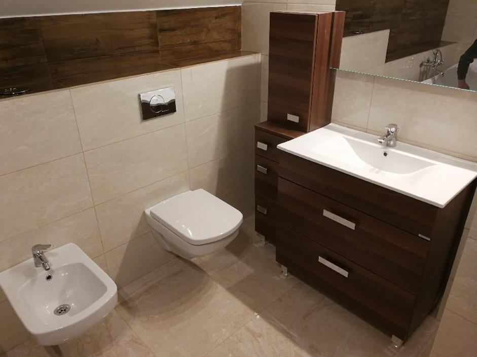 Nowoczesna łazienka z wiszącą toaletą, bidetem i szafką pod umywalkę w kolorze ciemnego drewna, wykończona beżowymi płytkami imitującymi kamień, z akcentem w postaci brązowego pasa płytek na górze...