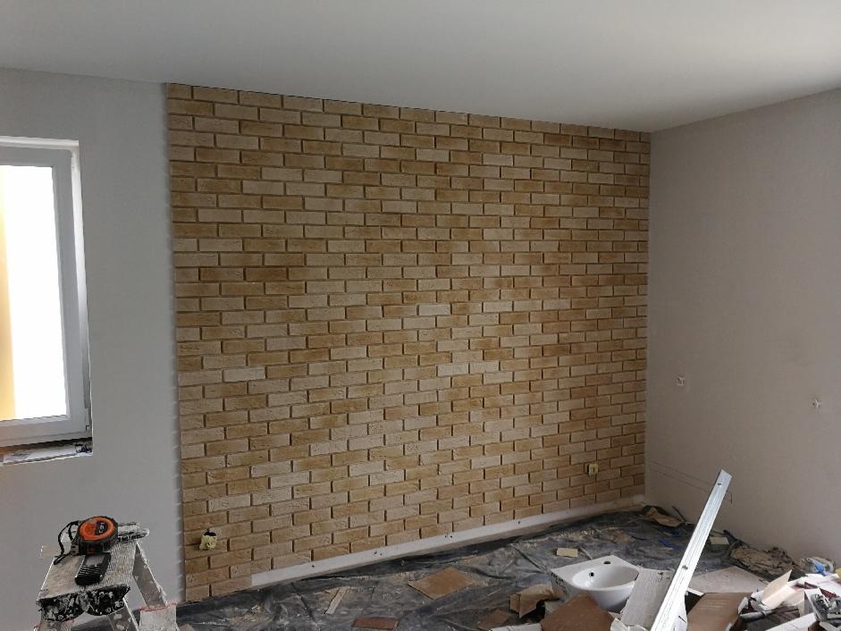 Ściana wykończona dekoracyjną cegłą w odcieniach beżu i brązu, widoczna z boku okna i częściowo zakryta folią ochronną na podłodze w trakcie remontu.