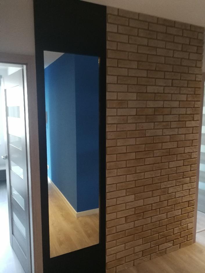 Ścianka działowa wykończona cegiełkami dekoracyjnymi w odcieniach beżu, przylegająca do czarnej ramy z lustrem, odbijającym niebieską ścianę i jasną podłogę.
