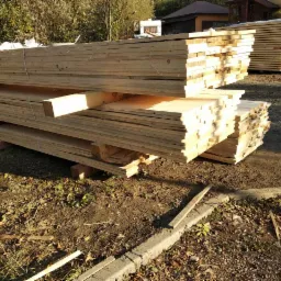 Stosy desek z jasnego drewna ułożone na paletach na zewnątrz, widoczne ślady jesiennych liści i resztek drewna na ziemi.