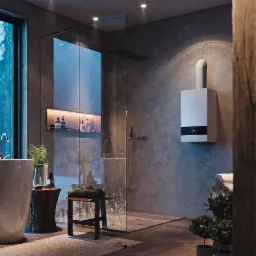 Nowoczesna łazienka z kabiną prysznicową typu walk-in i wiszącym piecem gazowym z białym kominem, oświetlona ciepłym światłem, z widocznym fragmentem drewnianej kolumny.