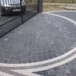 Ułożona z ciemnej kostki granitowej nawierzchnia podjazdu z dekoracyjnym, jasnym obramowaniem w kształcie łuku, widok zza otwartej, metalowej bramy.