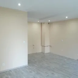 Pusty pokój po remoncie z szarymi panelami podłogowymi imitującymi drewno, beżowe ściany z widocznymi puszkami elektrycznymi i rurami, oraz białym sufitem z halogenami.