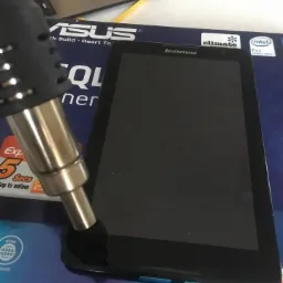 Tablet Lenovo na pudełku ASUS P5QL, obok lutownica na gorące powietrze i odseparowany ekran z notatką A8-50, sugerujące proces wymiany lub naprawy ekranu.