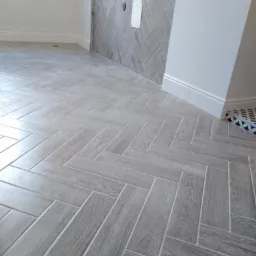 Jasnoszare płytki podłogowe ułożone w jodełkę, imitujące drewno, z fragmentem ściany wykończonej w identyczny sposób. Widoczny fragment listwy przypodłogowej.