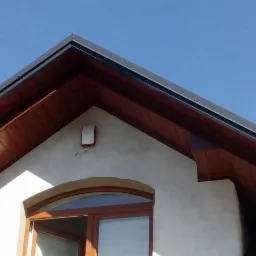 Fragment elewacji domu z drewnianą podbitką dachową, szarymi rynnami i oknem łukowym w stanie surowym.