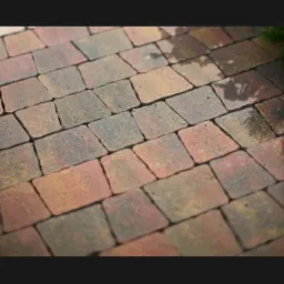 Ułożona nawierzchnia z wielokolorowej kostki brukowej w kształcie trapezu, z widocznymi fugami i fragmentem trawy w rogu.
