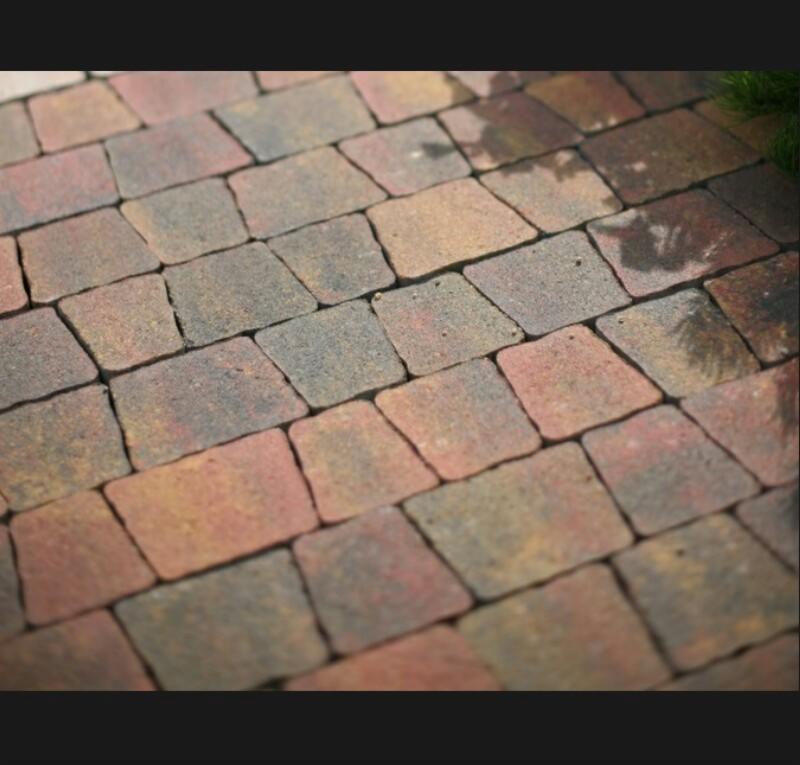 Ułożona nawierzchnia z wielokolorowej kostki brukowej w kształcie trapezu, z widocznymi fugami i fragmentem trawy w rogu.