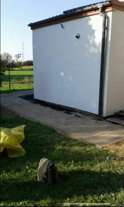 Biała ściana budynku z szarą kostką brukową wokół, widoczny worek z cementem na trawie i fragment żółtej plandeki