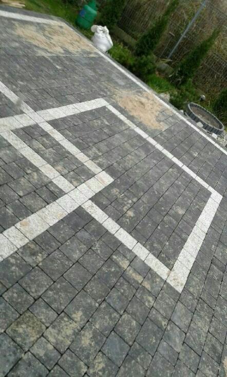 Ułożona nawierzchnia z kostki brukowej w odcieniach szarości, z białymi liniami tworzącymi geometryczny wzór, widoczne narzędzia i materiały budowlane w tle.