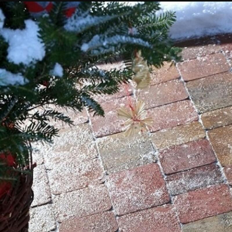 Kostka brukowa w odcieniach czerwieni i beżu, delikatnie przyprószona śniegiem, z fragmentem choinki w donicy i ozdobą świąteczną.