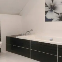 Nowoczesna łazienka z czarno-białymi płytkami, wisząca toaleta i wanna obudowana czarnymi płytkami, na ścianie grafika z motywem kwiatowym.