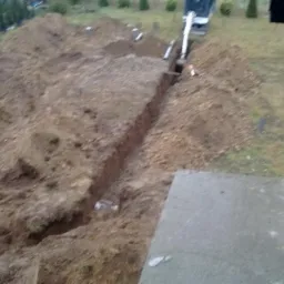 Wąski wykop w ziemi, wykonany przy użyciu minikoparki, widoczny z góry, z zielonym trawnikiem w tle i fragmentem betonowej konstrukcji w prawym dolnym rogu.