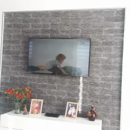 Ściana akcentowa z szarej imitacji kamienia, podwieszony telewizor, biała szafka RTV z dekoracjami i kwiatami w salonie.