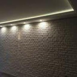 Igbud - Ściana wyłożona cegiełkami dekoracyjnymi w odcieniach bieli i szarości, oświetlona od góry listwą LED w suficie, tworząca nowoczesny efekt wizualny.