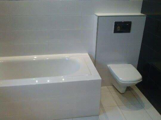 Biała, minimalistyczna łazienka z wanną obudowaną płytkami, podwieszanym WC z czarnym przyciskiem spłukującym i czarną ścianą z płytek.