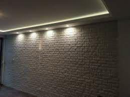 Ściana wyłożona cegiełkami dekoracyjnymi w odcieniach bieli i szarości, oświetlona od góry listwą LED w suficie, tworząca nowoczesny efekt wizualny.