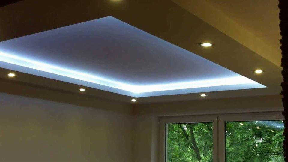 Podwieszany sufit z oświetleniem LED i punktowymi lampami halogenowymi, widok z dołu, jasne ściany i okno z widokiem na zieleń.