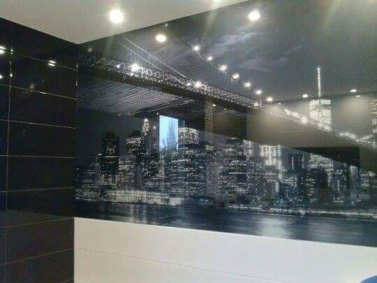 Czarno-biała fototapeta z motywem panoramy Nowego Jorku, w tym mostu Brooklyńskiego i Empire State Building, zamontowana w łazience nad białą zabudową, obok ściany wyłożonej czarnymi, błyszczącymi...