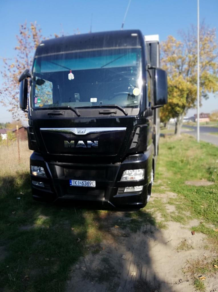 Czarny ciągnik siodłowy marki MAN z numerem rejestracyjnym TKI 6369C, zaparkowany na trawiastym poboczu w słoneczny dzień.