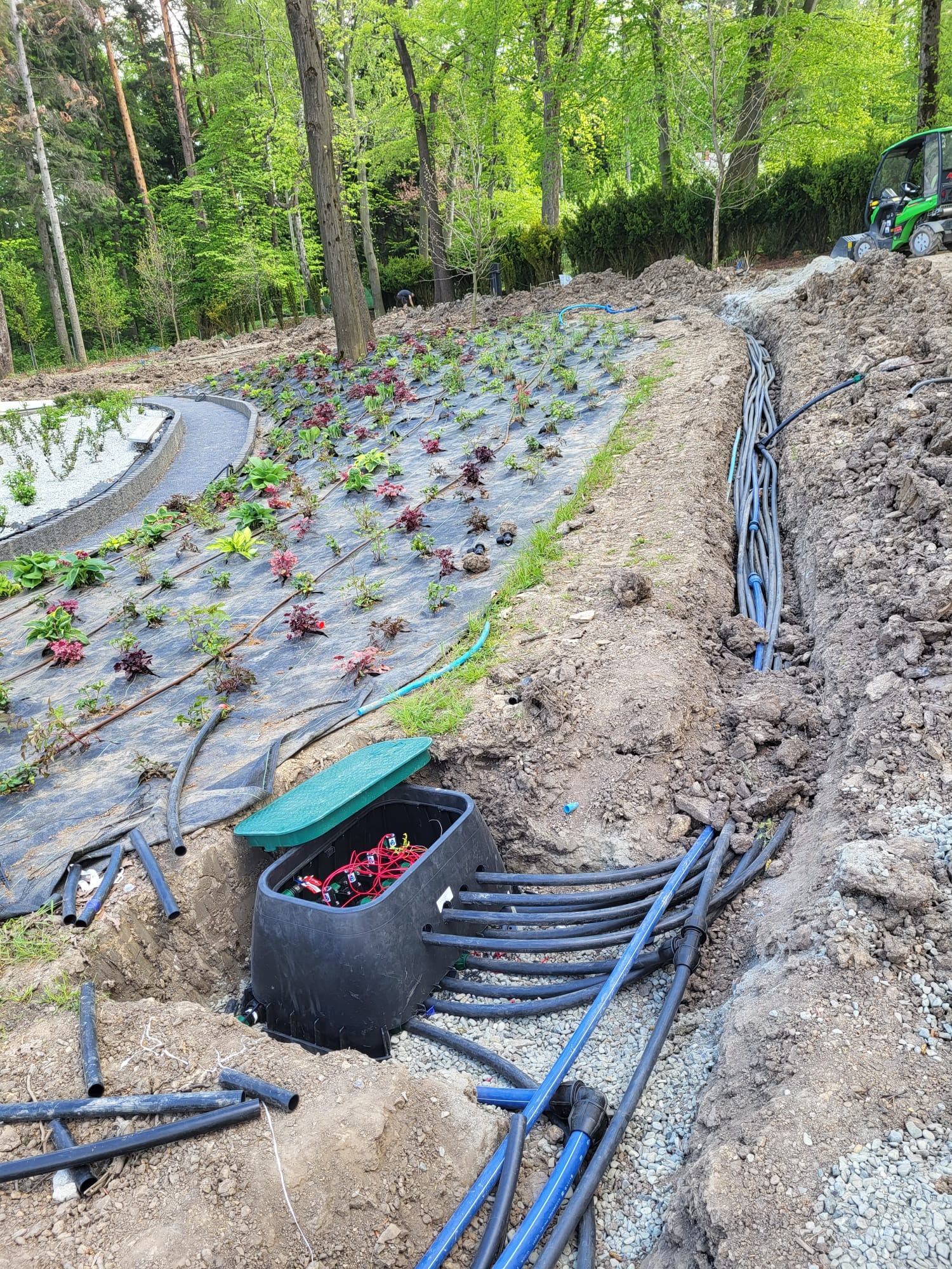 Instalacja systemu nawadniania w ogrodzie: widok na rozłożone węże nawadniające, czarną skrzynkę sterującą z otwartą zieloną pokrywą oraz wykopany rów z ułożonymi rurami doprowadzającymi wodę,...