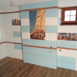 Wykończona łazienka z motywem marynistycznym: biało-błękitne płytki z dekoracyjnym wzorem żaglowca i drewnianym akcentem, brązowa drewniana podłoga, drewniane okno, surowy sufit z wiszącą żarówką.