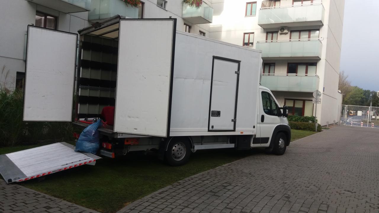 Biała furgonetka z otwartymi tylnymi drzwiami i rampą rozładowczą, zaparkowana na kostce brukowej przed nowoczesnym budynkiem mieszkalnym z balkonami.