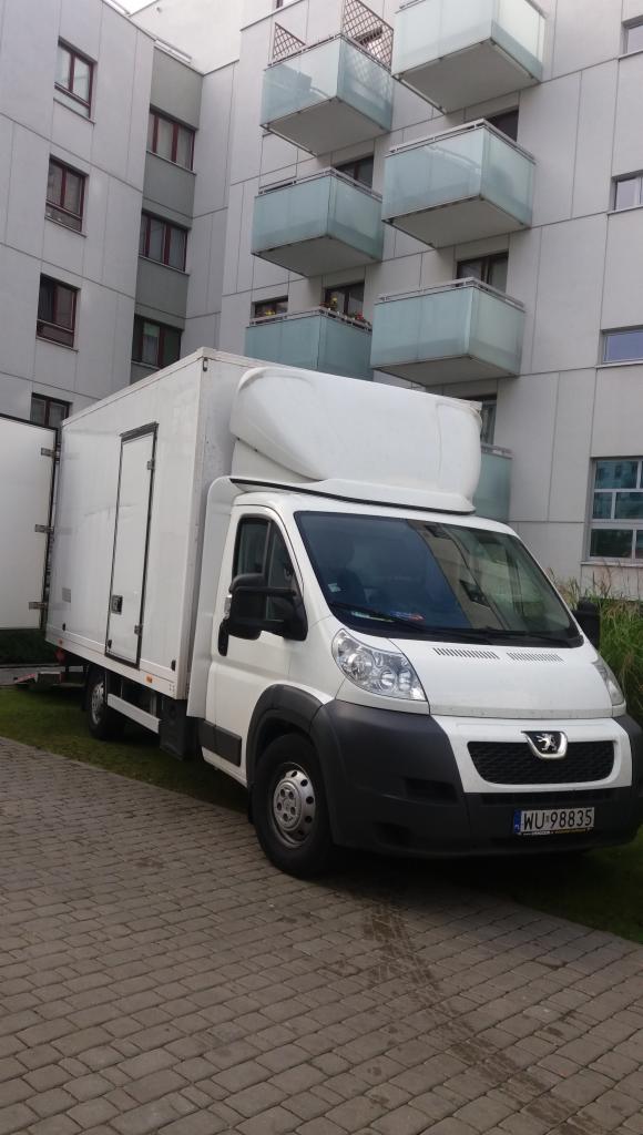 Biała furgonetka z nadbudową zaparkowana przed nowoczesnym budynkiem mieszkalnym z balkonami, widoczna tablica rejestracyjna.