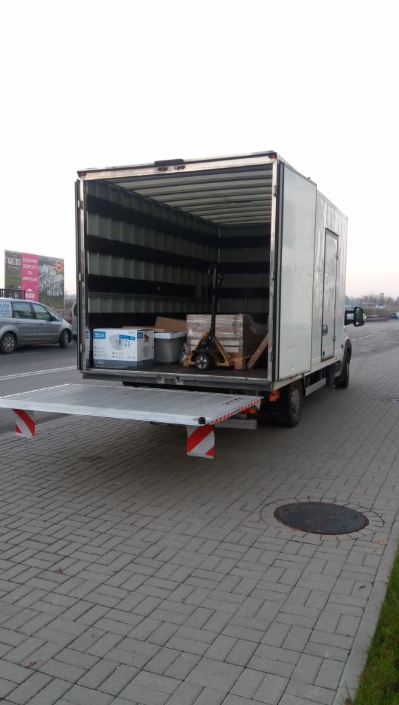 Otwarta rampa załadunkowa białej ciężarówki z częściowo załadowanym towarem: pudła, wiadro i paleta z płytkami, w tle wózek paletowy.