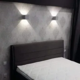 Sypialnia z łóżkiem z ciemnym wezgłowiem, ściany w jasne wzory, oświetlenie punktowe w postaci dwóch kwadratowych lamp ściennych, ciemny pas dekoracyjny pod sufitem.