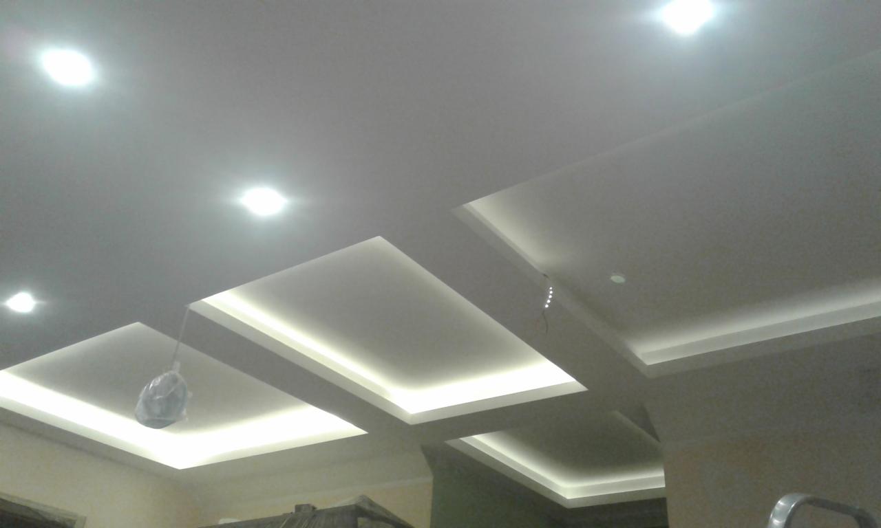 Nowoczesny sufit podwieszany z oświetleniem LED w kształcie prostokątów, widoczne przewody i folia ochronna na lampie, jasne światło, ujęcie z dołu.