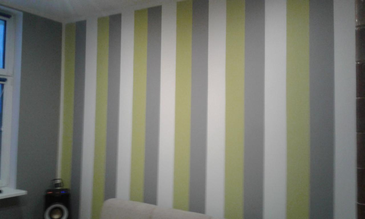 Ściana w pionowe pasy w odcieniach szarości, bieli i oliwkowej zieleni, z widocznym fragmentem okna po lewej stronie i głośnikiem na parapecie.