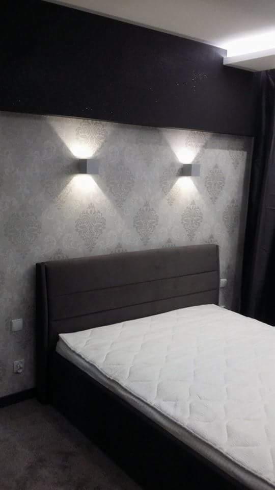 Sypialnia z łóżkiem z ciemnym wezgłowiem, ściany w jasne wzory, oświetlenie punktowe w postaci dwóch kwadratowych lamp ściennych, ciemny pas dekoracyjny pod sufitem.