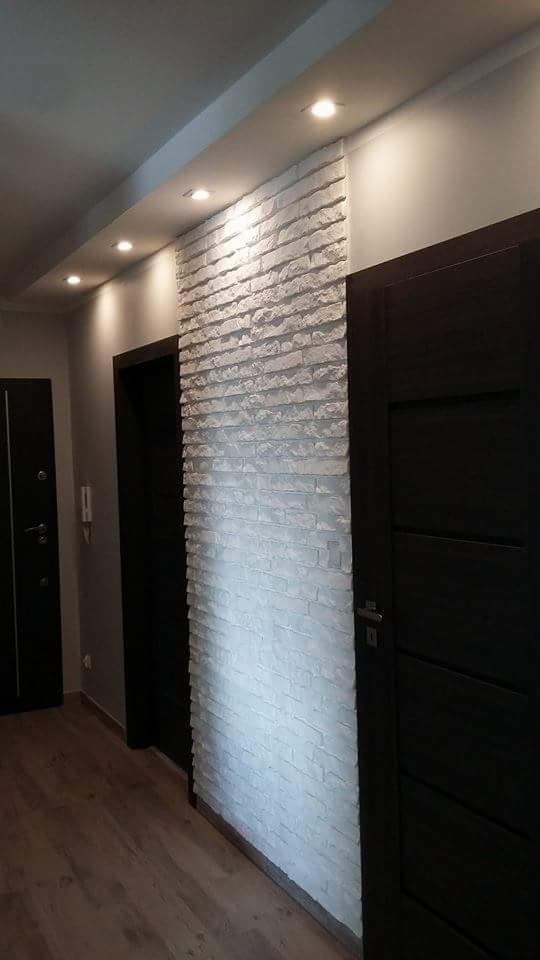 Ściana w korytarzu wyłożona dekoracyjną, białą cegłą, oświetlona punktowymi lampami sufitowymi, sąsiadująca z ciemnymi drzwiami.