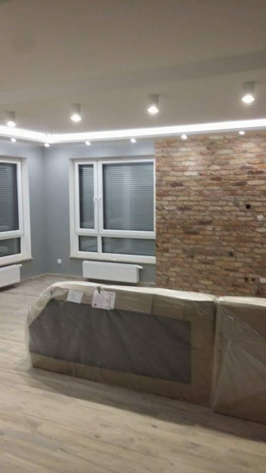 Wnętrze pokoju po remoncie z wyeksponowaną ścianą z cegły, parkietem i oknami z żaluzjami, częściowo zasłoniętym meblem w folii ochronnej.