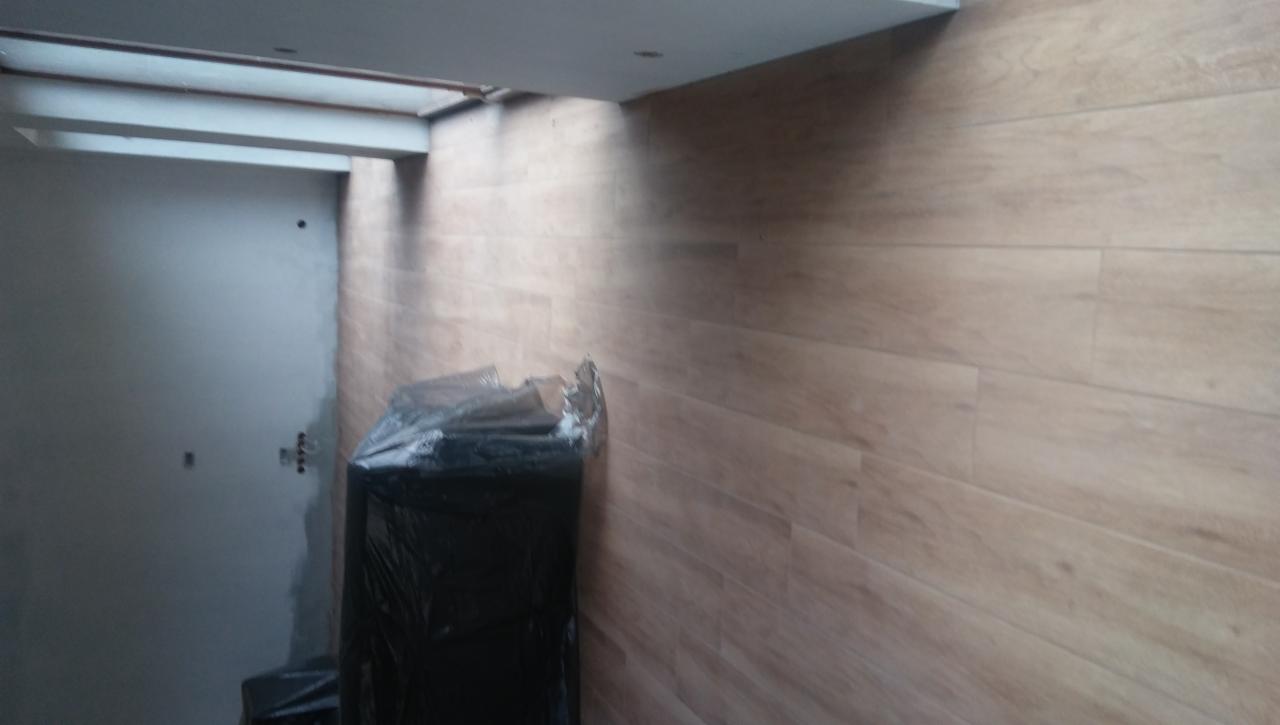 Ściana wykończona panelami imitującymi drewno, z widocznym narożnikiem sufitu i fragmentem szarej, surowej ściany w tle, przy której stoi czarny worek na śmieci.