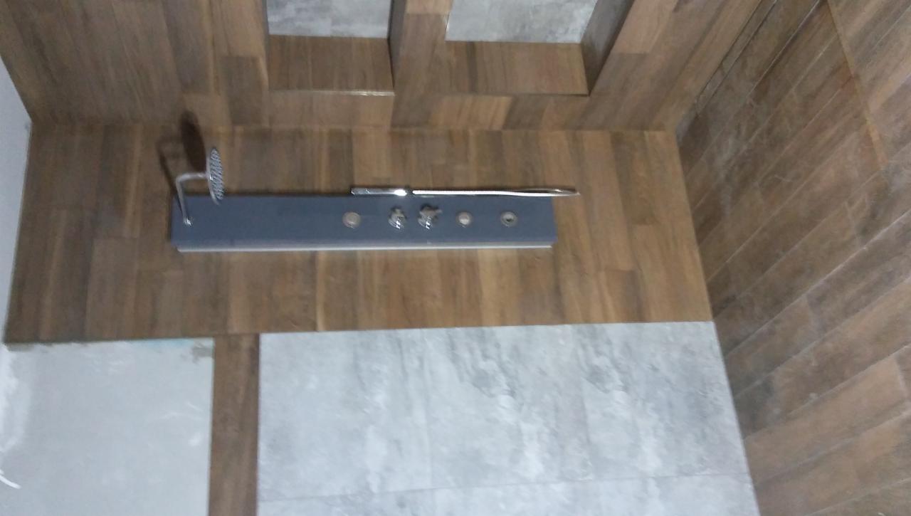 Montaż panelu prysznicowego z deszczownicą na brązowej ścianie imitującej drewno, widoczne płytki podłogowe w odcieniach szarości.