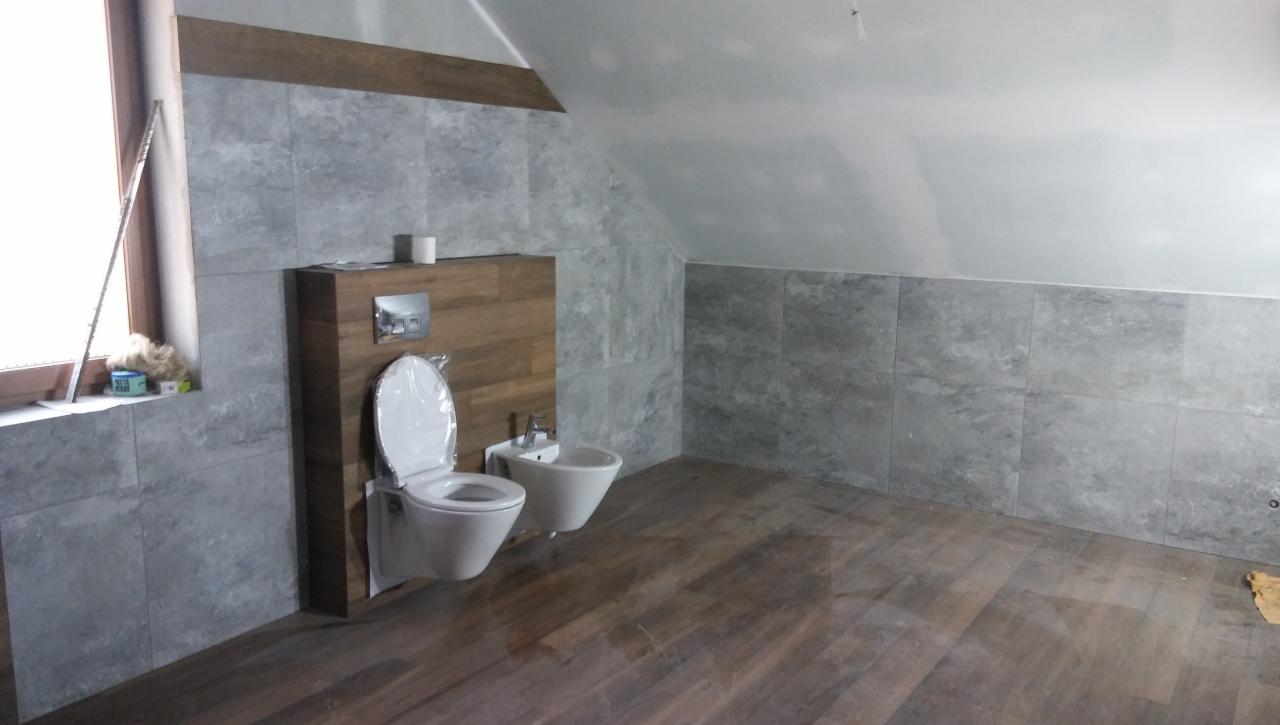 Wykończona łazienka na poddaszu z szarymi płytkami imitującymi kamień, drewnopodobną zabudową WC i podłogą, białą miską WC w folii ochronnej i bidetem.