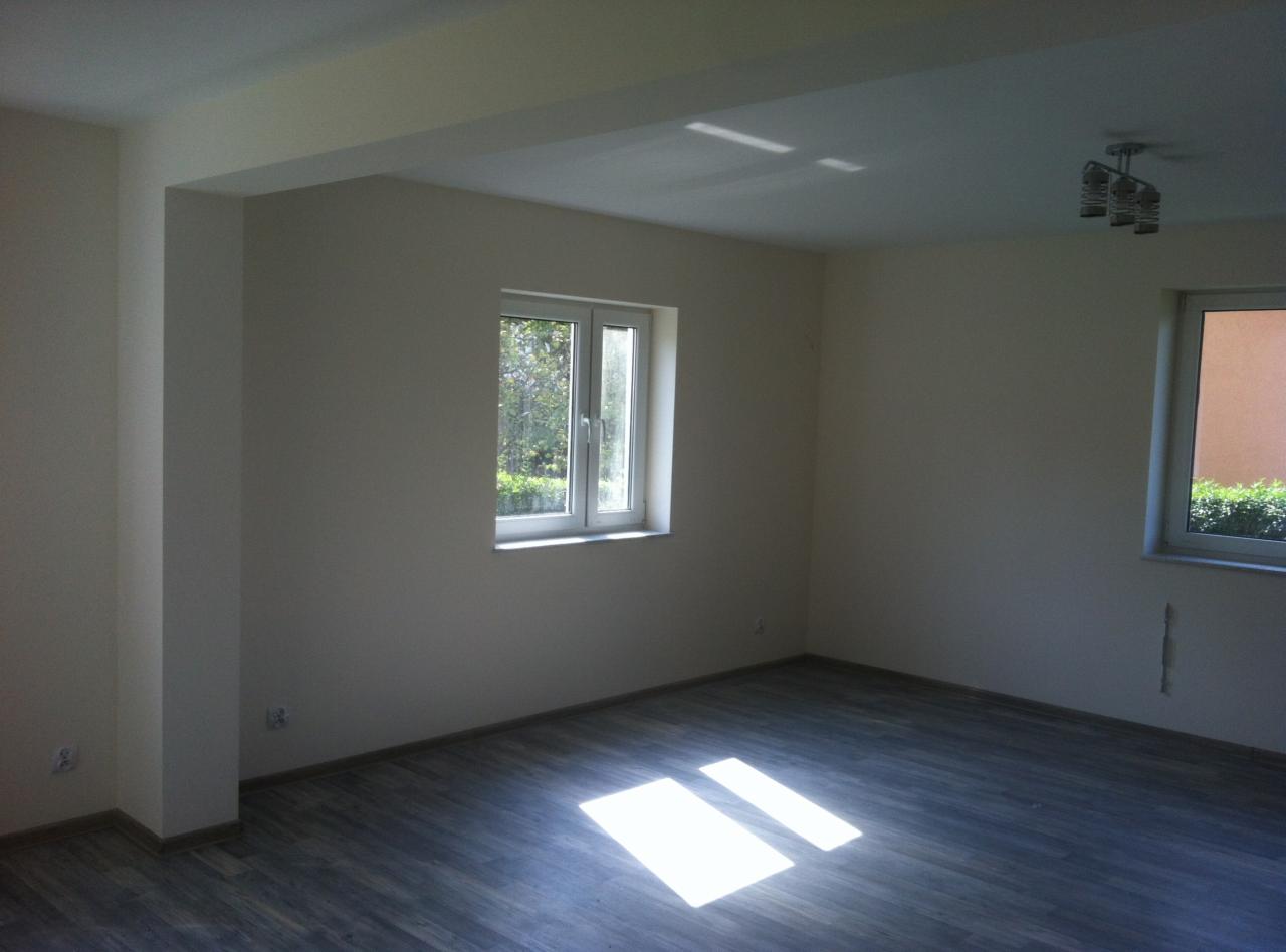 Pomalowane na jasny kolor wnętrze pokoju z parkietem, widoczne dwa okna z widokiem na zieleń na zewnątrz oraz lampa sufitowa.